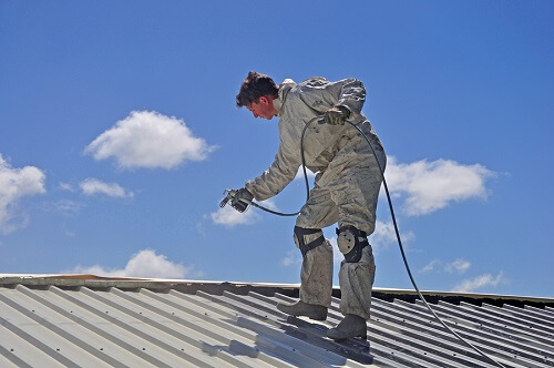 Roof painter wearing safety gear and spraying a metal roof.